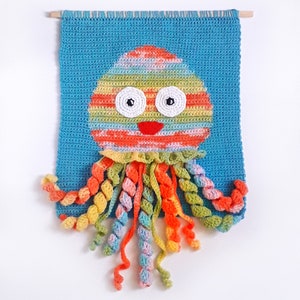 Puede incluir: Un tapiz de pared de ganchillo con un pulpo colorido con una cabeza a rayas y tent&aacute;culos rizados. El pulpo tiene una boca roja y ojos blancos.