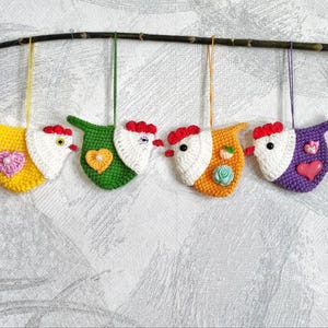 May include: Five crocheted chicken ornaments hanging from a branch. The chickens are yellow, green, orange, purple, and white with red beaks and white eyes. Each chicken has a small heart-shaped button on its chest.