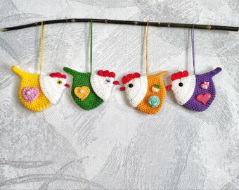 May include: Five crocheted chicken ornaments hanging from a branch. The chickens are yellow, green, orange, purple, and white with red beaks and white eyes. Each chicken has a small heart-shaped button on its chest.
