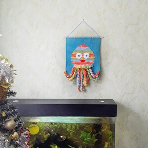 Puede incluir: Un tapiz de pared de ganchillo de color azul y naranja con ojos blancos y una sonrisa roja. El pulpo est&aacute; colgado de una cuerda en una pared blanca. En primer plano, hay un tanque de peces negro con plantas y un pez.