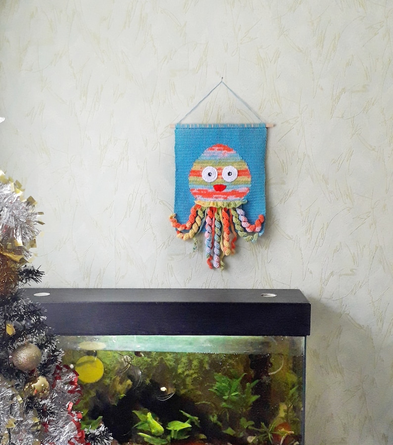 Puede incluir: Un tapiz de pared de ganchillo de color azul y naranja con ojos blancos y una sonrisa roja. El pulpo est&aacute; colgado de una cuerda en una pared blanca. En primer plano, hay un tanque de peces negro con plantas y un pez.