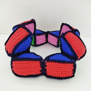 May include: A colorful, crocheted necklace featuring a geometric design. The necklace is composed of connected rectangular shapes in red, blue, and pink, outlined in black. A touch of green is visible. The craftwork is displayed against a white background.