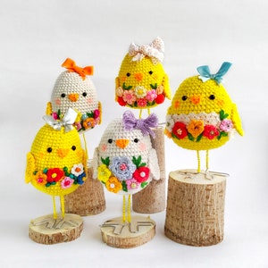 Op de afbeelding: Vijf gehaakte kuikens met bloemmotieven staan op houten bases. Elk kuiken heeft een ander kleurenschema en bloemmotief.