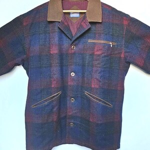 May include: A plaid jacket in shades of blue, burgundy, and green, with a brown collar and pocket trim. The jacket has a button-up front and a zippered chest pocket. The jacket is hanging on a wooden hanger.