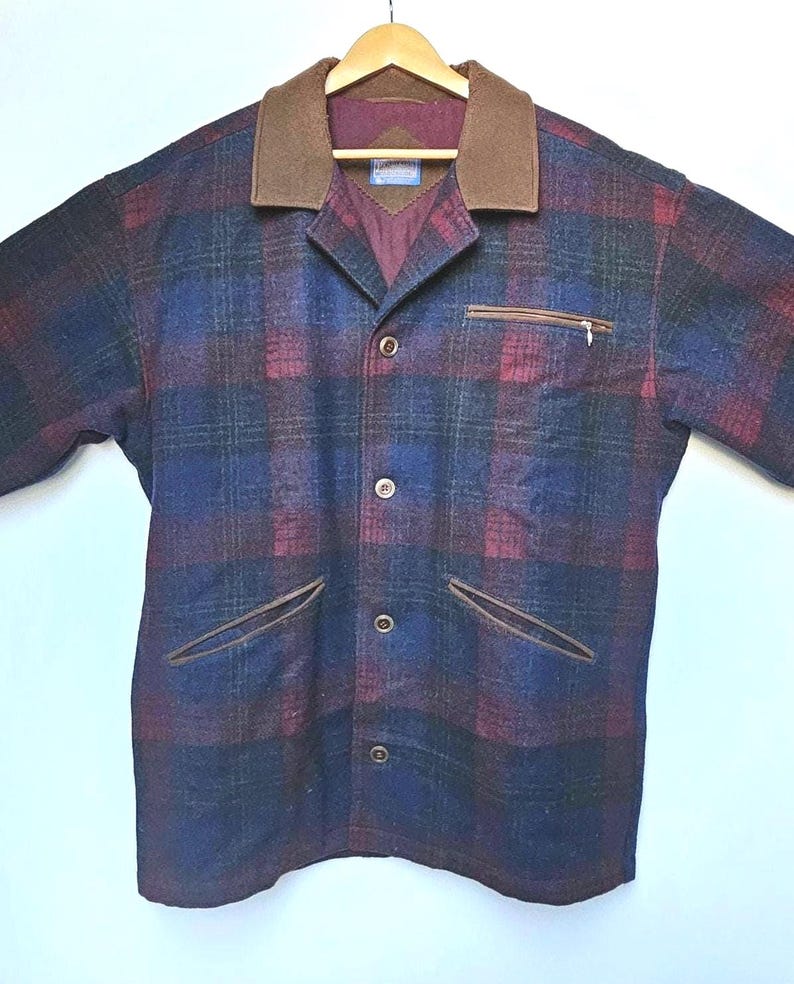 May include: A plaid jacket in shades of blue, burgundy, and green, with a brown collar and pocket trim. The jacket has a button-up front and a zippered chest pocket. The jacket is hanging on a wooden hanger.