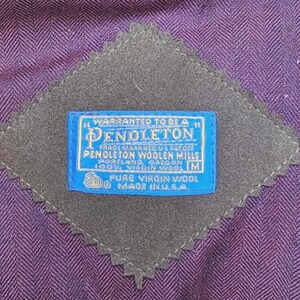 May include: Close-up of a purple herringbone fabric with a gray, diamond-shaped patch. A blue label with white text reads "Pendleton Woolen Mills, Portland, Oregon, Pure Virgin Wool, Made in U.S.A."