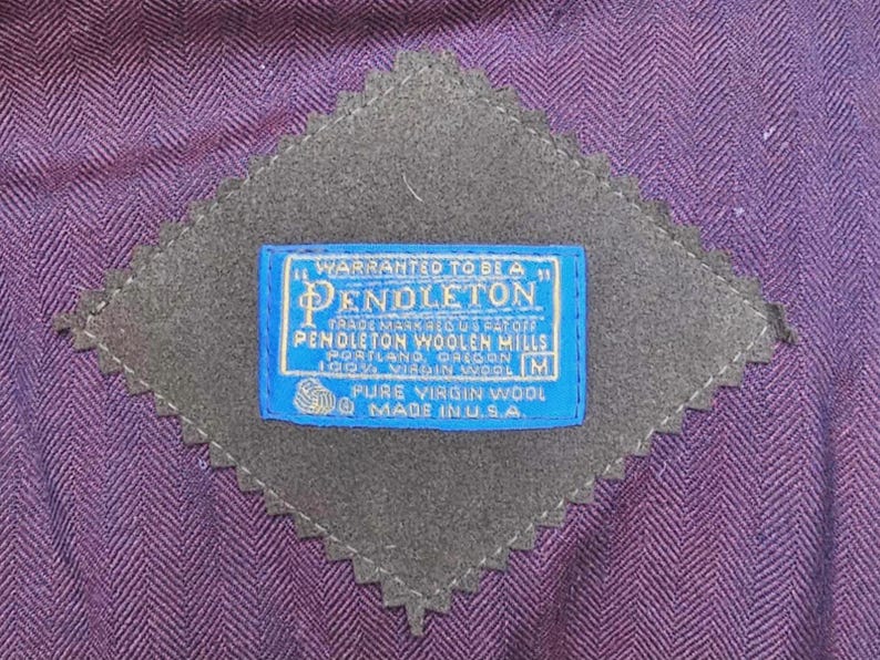 May include: Close-up of a purple herringbone fabric with a gray, diamond-shaped patch. A blue label with white text reads "Pendleton Woolen Mills, Portland, Oregon, Pure Virgin Wool, Made in U.S.A."