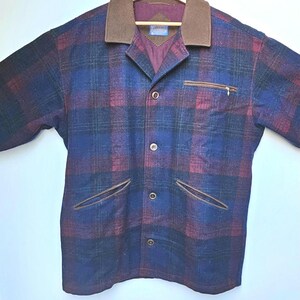 May include: A plaid jacket in shades of blue, burgundy, and green, with a brown collar and pocket trim. The jacket has a button-up front, a chest pocket with a zipper, and two front pockets. It is hanging on a wooden hanger.