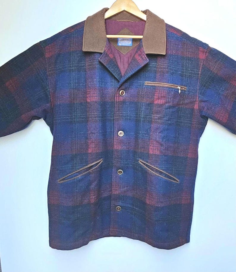 May include: A plaid jacket in shades of blue, burgundy, and green, with a brown collar and pocket trim. The jacket has a button-up front, a chest pocket with a zipper, and two front pockets. It is hanging on a wooden hanger.