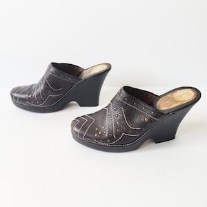 May include: A pair of brown leather wedge clogs with gold-toned studs and stitching. The clogs have a closed toe and a slip-on design.