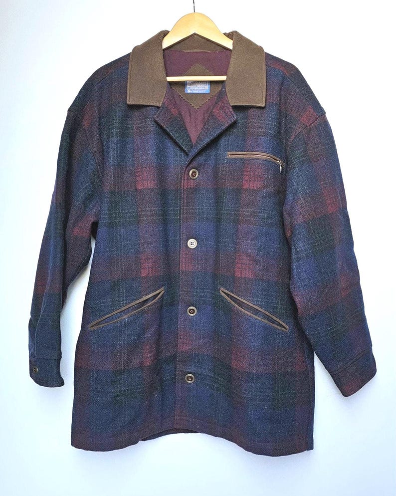 May include: A plaid wool jacket in shades of blue, burgundy, and green, featuring a brown collar and pocket trim. The jacket has a button-up front, a zippered chest pocket, and two front pockets. It is hanging on a wooden hanger.