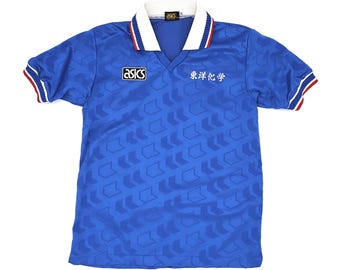 Puede incluir: Camiseta deportiva azul con cuello blanco y ribetes rojos. La camiseta presenta el logo "asics" y texto japonés. La tela tiene un patrón de flechas repetidas. Una camiseta deportiva vintage.