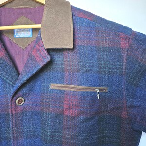 May include: A blue, red, and green plaid jacket with a brown collar and a zippered chest pocket. The jacket has brown buttons and a label with text. The jacket is hanging on a wooden hanger.