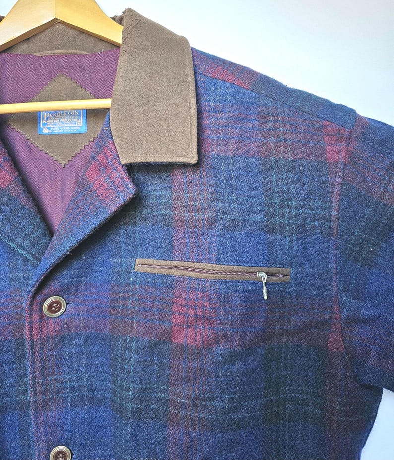 May include: A blue, red, and green plaid jacket with a brown collar and a zippered chest pocket. The jacket has brown buttons and a label with text. The jacket is hanging on a wooden hanger.