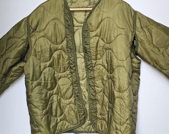 Puede incluir: Chaqueta acolchada verde oliva con diseño de cuello en V. La chaqueta está colgada de una percha de madera. La chaqueta tiene un patrón acolchado en el cuerpo y las mangas.