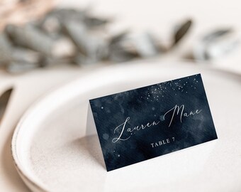 May include: A navy blue place card with the name "Lauren Mane" in elegant script. The card has a watercolor effect with white speckles, and is set on a white plate with gold cutlery. The words "TABLE 7" are also printed on the card.