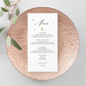 May include: A wedding menu card with a gold celestial design, featuring a crescent moon and stars. The menu lists canapés, hors d'oeuvres, entree, and dessert options. The card is on a rose gold plate, with eucalyptus leaves in the background.