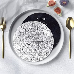 May include: A celestial-themed table setting featuring a white plate with a black and white star chart design. A gold fork and spoon flank the plate, with a menu card that reads "MENU". Fresh figs add a touch of color.