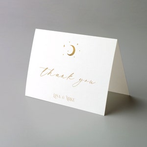 May include: A folded white card with gold foil lettering. The card reads "thank you" in a script font, with "LISA & MIKE" below. A gold crescent moon and stars are at the top.