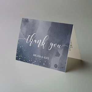 May include: A folded thank you card with a watercolor design in shades of blue and gray. The words "thank you" are written in elegant white script, with "MILANA & ALEX" below. Silver glitter accents the design.