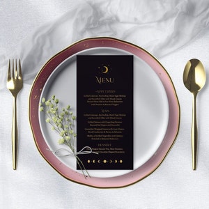 May include: A formal place setting with a black menu card featuring gold text and moon phase graphics. The plate is white with a pink and gold rim. A gold fork and spoon flank the plate, and a small bouquet of white flowers rests on the plate.