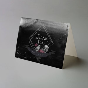 May include: A folded thank you card with a dark, watercolor-style background. The card features a geometric frame with the words "Thank You" in elegant script. Below, a banner displays the names "Amelia & Victor" alongside illustrations of an owl, skull, and flowers.