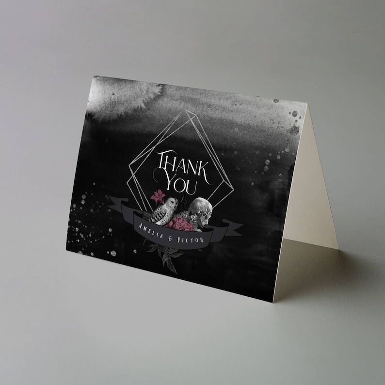 May include: A folded thank you card with a dark, watercolor-style background. The card features a geometric frame with the words "Thank You" in elegant script. Below, a banner displays the names "Amelia & Victor" alongside illustrations of an owl, skull, and flowers.