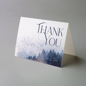 May include: A folded thank you card with a forest scene in shades of blue and gray. The words "THANK YOU" are written in elegant navy blue script. Small birds fly across the sky above the trees. The names "Anna & Zeger" are written in script at the bottom.