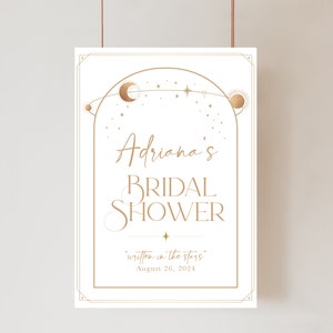 May include: A white sign with gold celestial design elements, including a crescent moon, stars, and a sun. The sign reads "Adriana's BRIDAL SHOWER" with the phrase "written in the stars" and the date August 26, 2024.