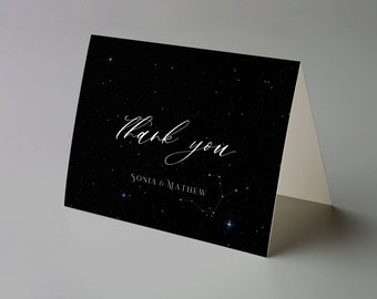 Puede incluir: Tarjeta de agradecimiento plegada con un fondo negro con diseño de cielo estrellado. Las palabras "Thank you" están escritas en elegante escritura blanca, con "Sonia & Matthew" debajo. También se ve una constelación.