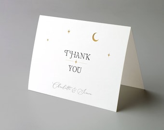 Puede incluir: Tarjeta de agradecimiento blanca plegada con detalles celestiales dorados. La tarjeta presenta las palabras "THANK YOU" en una fuente serif, con los nombres "Charlotte & Simon" debajo. Estrellas doradas y una luna creciente decoran la parte superior.