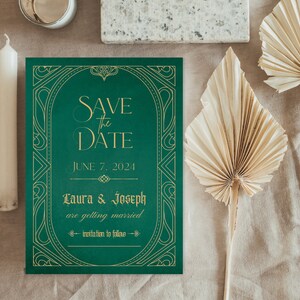 May include: Emerald green "Save the Date" card with gold Art Deco style lettering and design. The card reads "Save the Date" and "June 7, 2024" with the names "Laura & Joseph" and "invitation to follow". A white candle and dried palm leaves are also visible.