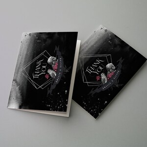 May include: Two folded thank you cards with a black watercolor background. The cards feature a geometric design with the words "THANK YOU" in white. Below the text is a banner with a skull, owl, and flowers. The banner reads "AMELIA & VICTOR".