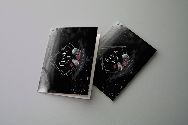 May include: Two folded thank you cards with a black watercolor background. The cards feature a geometric design with the words "THANK YOU" in white. Below the text is a banner with a skull, owl, and flowers. The banner reads "AMELIA & VICTOR".