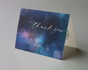 Puede incluir: Una tarjeta de felicitación plegada con un diseño cósmico. La parte delantera presenta una impresión de galaxia azul oscuro, púrpura y verde azulado con las palabras "thank you" en una elegante escritura blanca. El interior es de color beige claro.