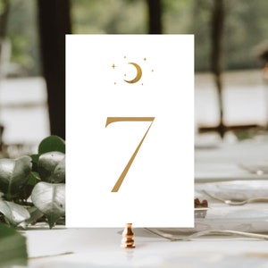 May include: A white table number card with a gold crescent moon and star design at the top. The number 7 is printed in gold. The card is held by a copper-colored clip, with a blurred background of greenery and a table setting.