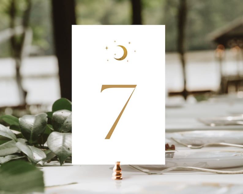 May include: A white table number card with a gold crescent moon and star design at the top. The number 7 is printed in gold. The card is held by a copper-colored clip, with a blurred background of greenery and a table setting.