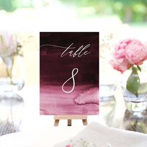 May include: A burgundy watercolor table number card with the word "table" in cursive and the number "8" in white. The card is displayed on a small wooden easel, with a blurred background of greenery and flowers.