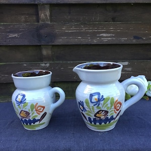 May include: Two white ceramic pitchers with floral designs in blue, orange, and green. The smaller pitcher has a handle on the side, while the larger pitcher has a handle on the top.