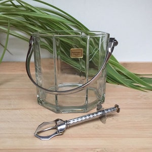 Puede incluir: Un cubo de hielo de vidrio transparente con un asa plateada y una pinza para hielo plateada. El cubo tiene una etiqueta que dice "Luminarc".