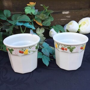 Peut inclure: Deux tasses en céramique blanche avec un motif floral représentant des poires jaunes, des fraises rouges et des feuilles vertes. Les tasses ont un fond blanc et un bord festonné.