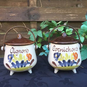 Puede incluir: Dos tarros de cerámica con tapas marrones y asas de metal. Los tarros están pintados con diseños florales y tienen las palabras "Oignons" y "Cornichons" escritas en ellos.