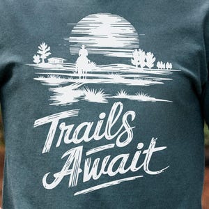 May include: Dark teal t-shirt with a white graphic of a cowboy on a horse in front of a sun and the words "Trails Await". The shirt is made of a soft material and is perfect for outdoor adventures.