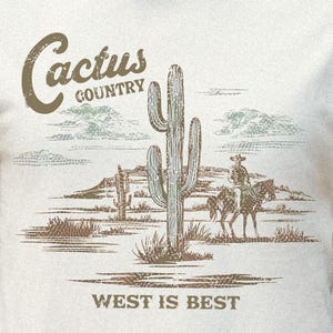 May include: Cream-colored t-shirt featuring a desert landscape design with a cactus, a cowboy on a horse, and the text "Cactus Country" and "West is Best". The graphic is in shades of brown and green.