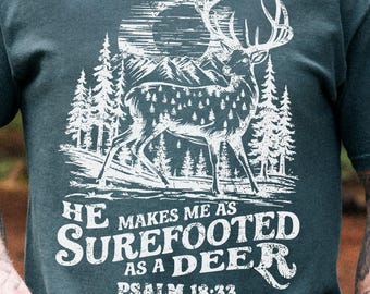 May include: Dark teal t-shirt featuring a white graphic of a deer with antlers, mountains, and pine trees. The text reads "He makes me as surefooted as a deer, Psalm 18:33."