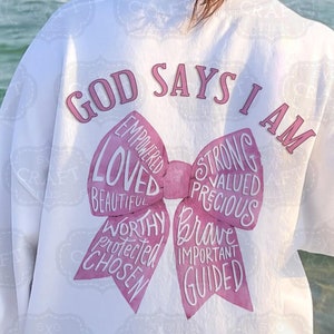 May include: White sweatshirt with the text "GOD SAYS I AM" and a pink bow graphic. The bow includes words like "Loved," "Strong," and "Brave." The sweatshirt is a casual style, suitable for everyday wear.
