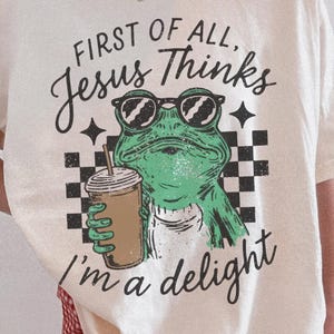 Funny Christian frog PNG holding iced coffee with First Of All Jesus Thinks I'm A Delight quote and checkerboard accents.