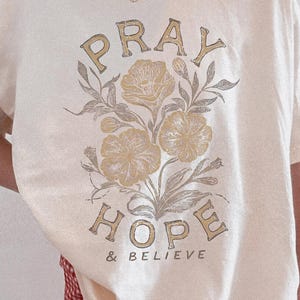 Może przedstawiać: Kremowa koszulka z napisem "PRAY HOPE & BELIEVE" w stylu vintage, z ilustracjami kwiatowymi. Modelka ma na sobie czerwono-białe szorty w kratę.