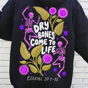 May include: A black hooded sweatshirt featuring a back graphic of two pink skeletons dancing among olive green leaves and bright purple flowers. The text reads "DRY BONES COME TO LIFE" in white letters, with "EZEKIEL 37:1-10" printed below in white.