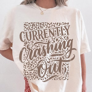 Puede incluir: Una camiseta de manga corta color blanco hueso con un gráfico rectangular de estampado de leopardo en el pecho. El texto "CURRENTLY" aparece en una fuente sans-serif negrita sobre el texto "crashing out" en color marrón oscuro.